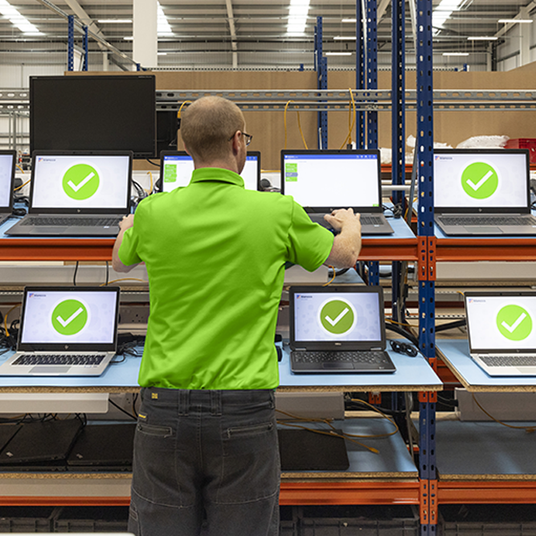 A technician standing in front of shelves holding laptops that show big green circles and white ticks as he runs quality control tests.