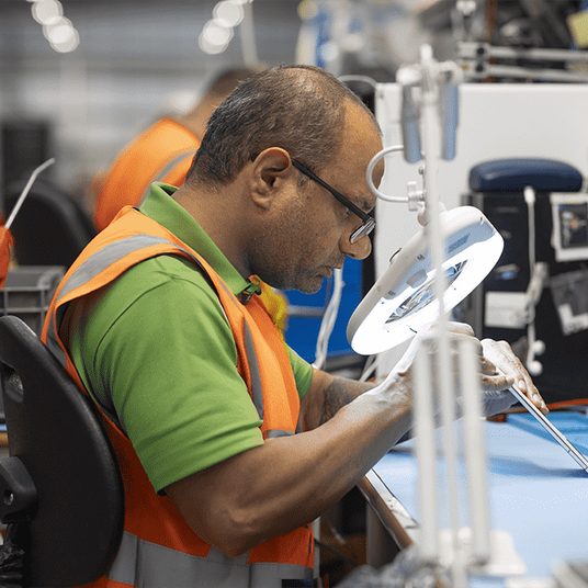 a technician wearing a high visibility waistcoat sits at a worktable and looks through a magnifying glass whilst repairing a device